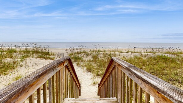 Une promenade en bois qui mène à une plage bordée de dunes de sable et d’une végétation clairsemée.