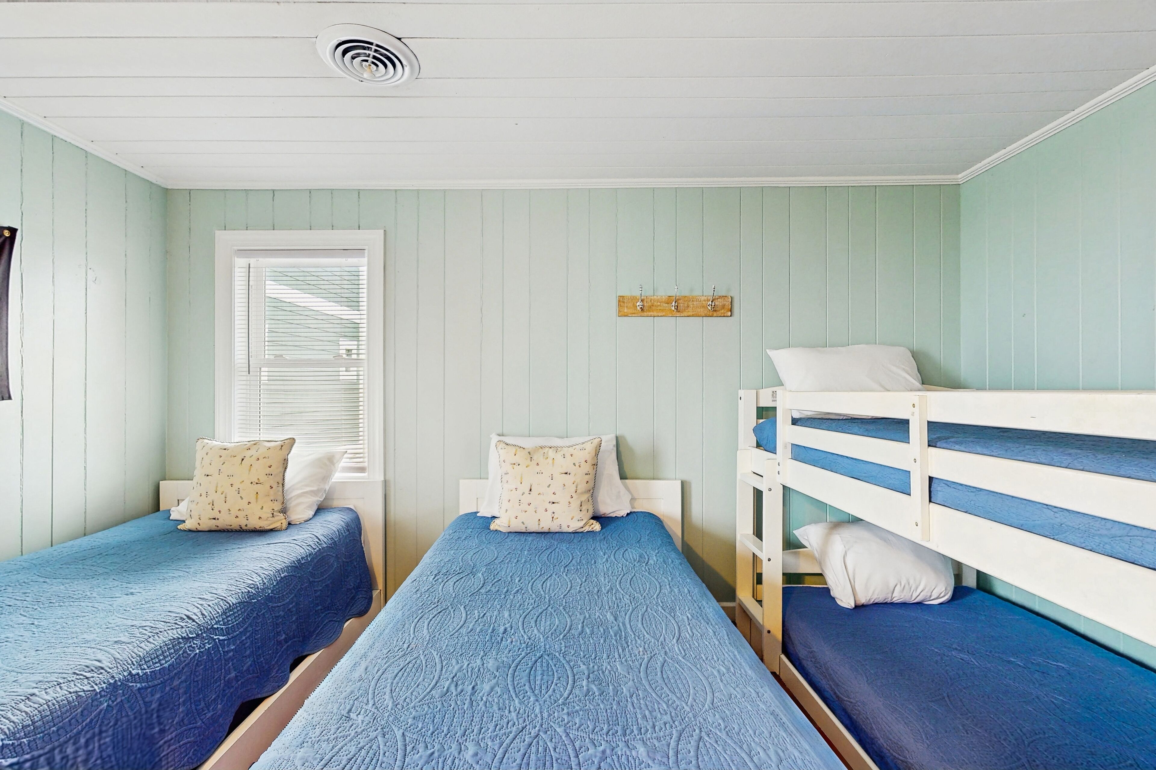 Una habitación con dos camas individuales, cada una con colcha y almohadas azules, cabecero blanco y ventana con persianas.