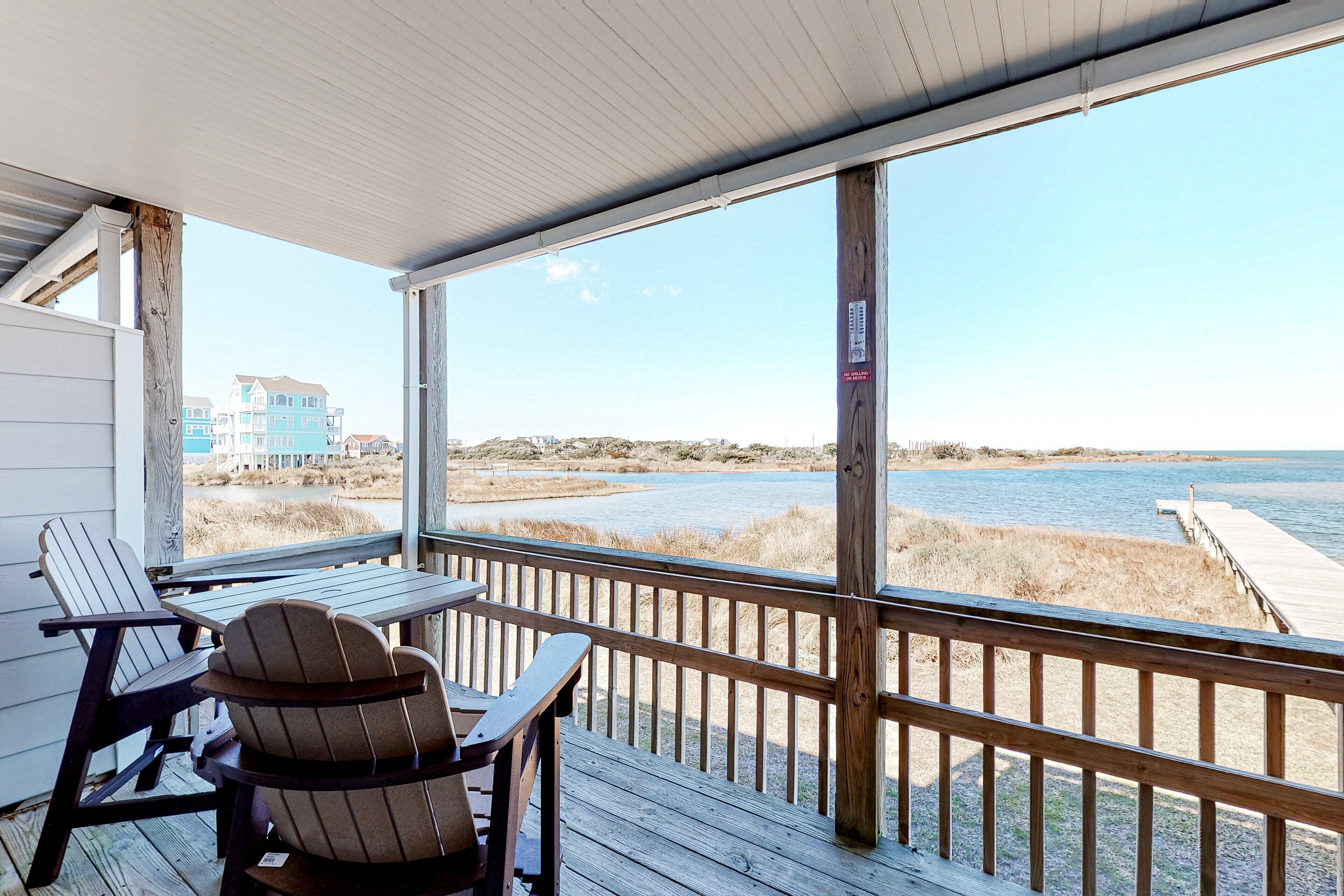 A wooden deck with two Adirondack chairs and a table, overlooking a beach and a building in the distance.