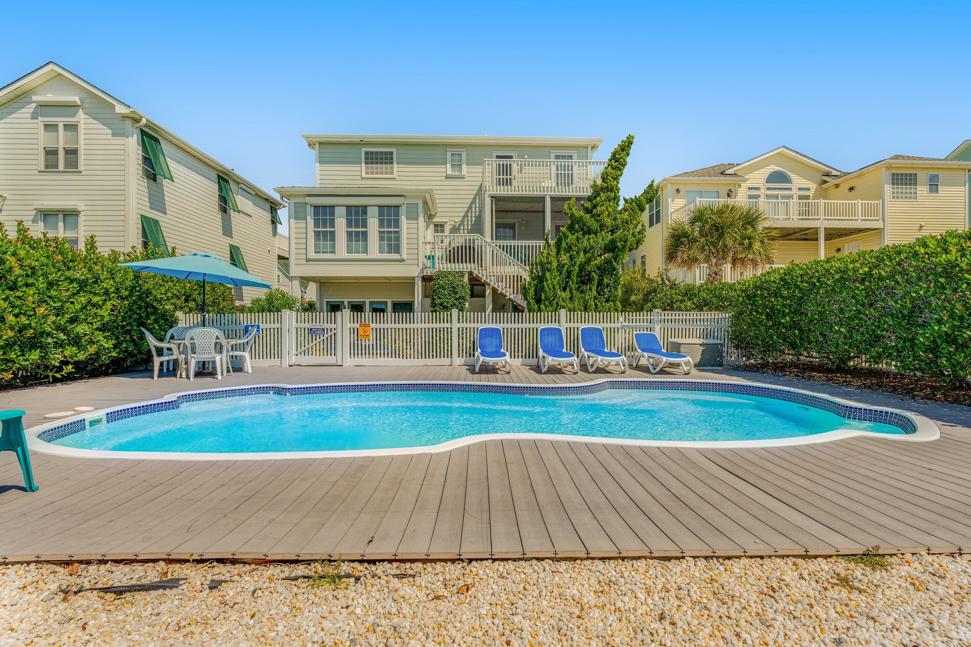 A swimming pool with a wooden deck, surrounded by a white picket fence, and adjacent to a multi-story building with balconies.