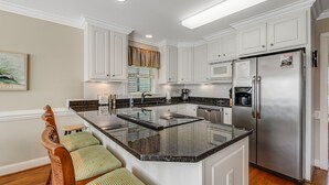 A modern kitchen with white cabinets, a stainless steel refrigerator, a granite countertop, and a wooden bench with patterned cushions. - Beautiful Waterway-View Home with Private Pool, Ping-Pong, Washer/Dryer & WiFi (Holden Beach)