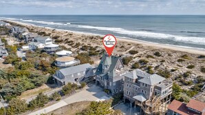 Una vista aerea di una grande casa a più piani con una torre imponente, situata vicino a una spiaggia, con un segnale rosso che indica Hatteras Realty.