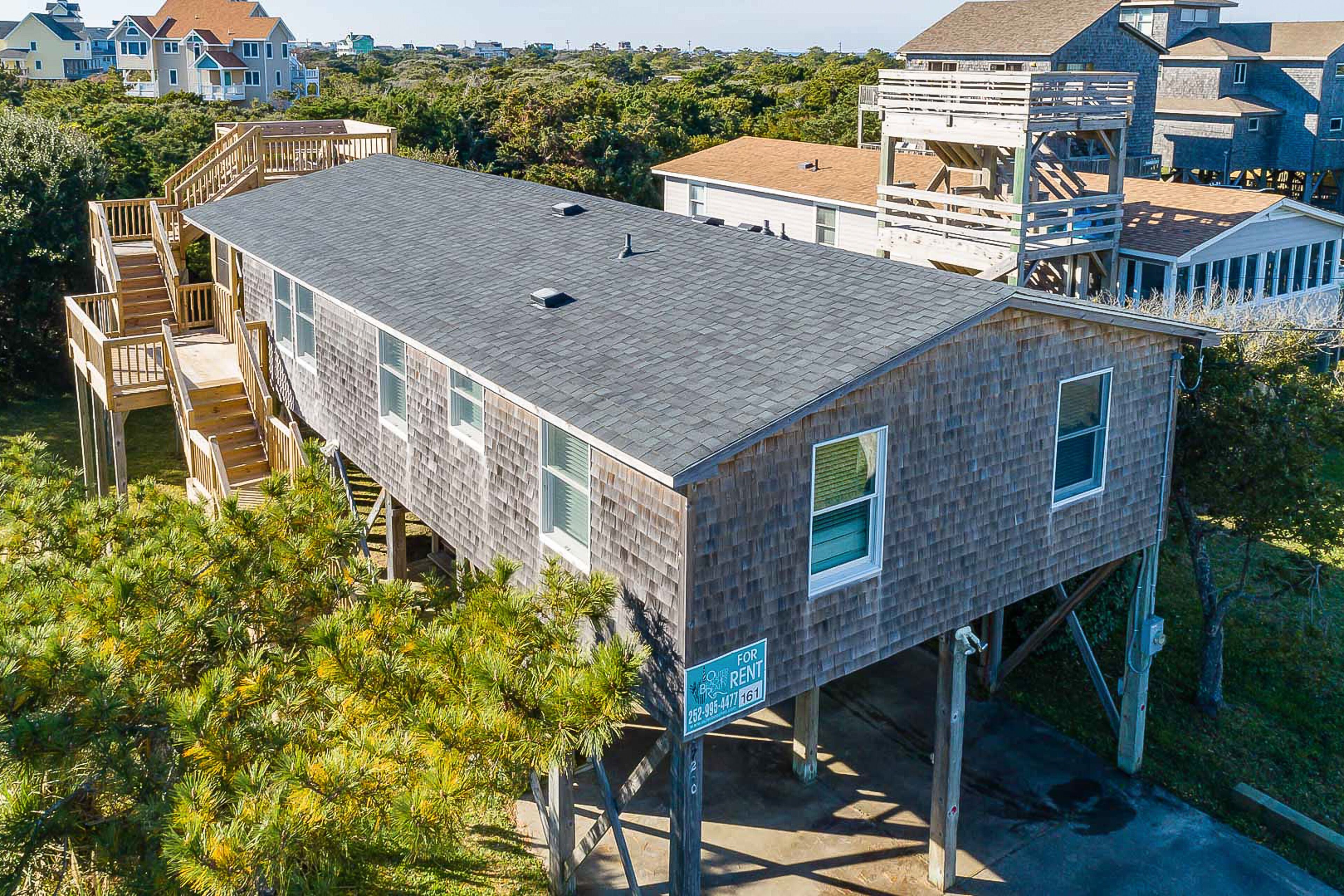 A two-story wooden house with a balcony, a 'For Rent' sign, and a view of surrounding houses and trees.