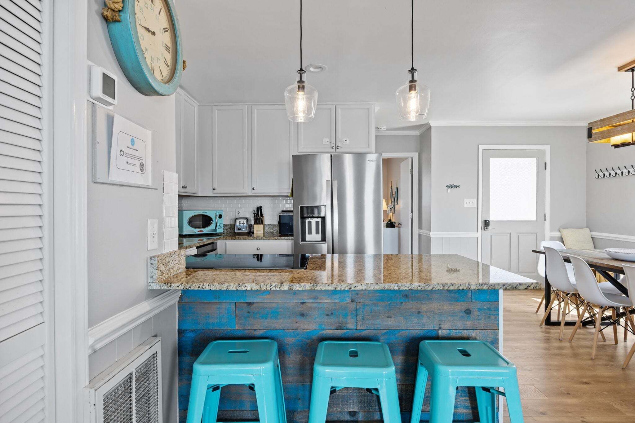 Une cuisine dotée d’un îlot central, d’appareils électroménagers en acier inoxydable, de luminaires suspendus et d’une horloge au mur.