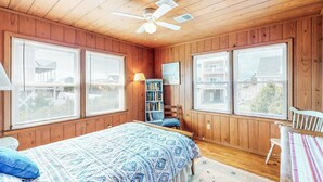 Une chambre avec des murs en bois, un ventilateur de plafond, un lit recouvert d’une courtepointe à motifs, une bibliothèque et de grandes fenêtres équipées de stores.