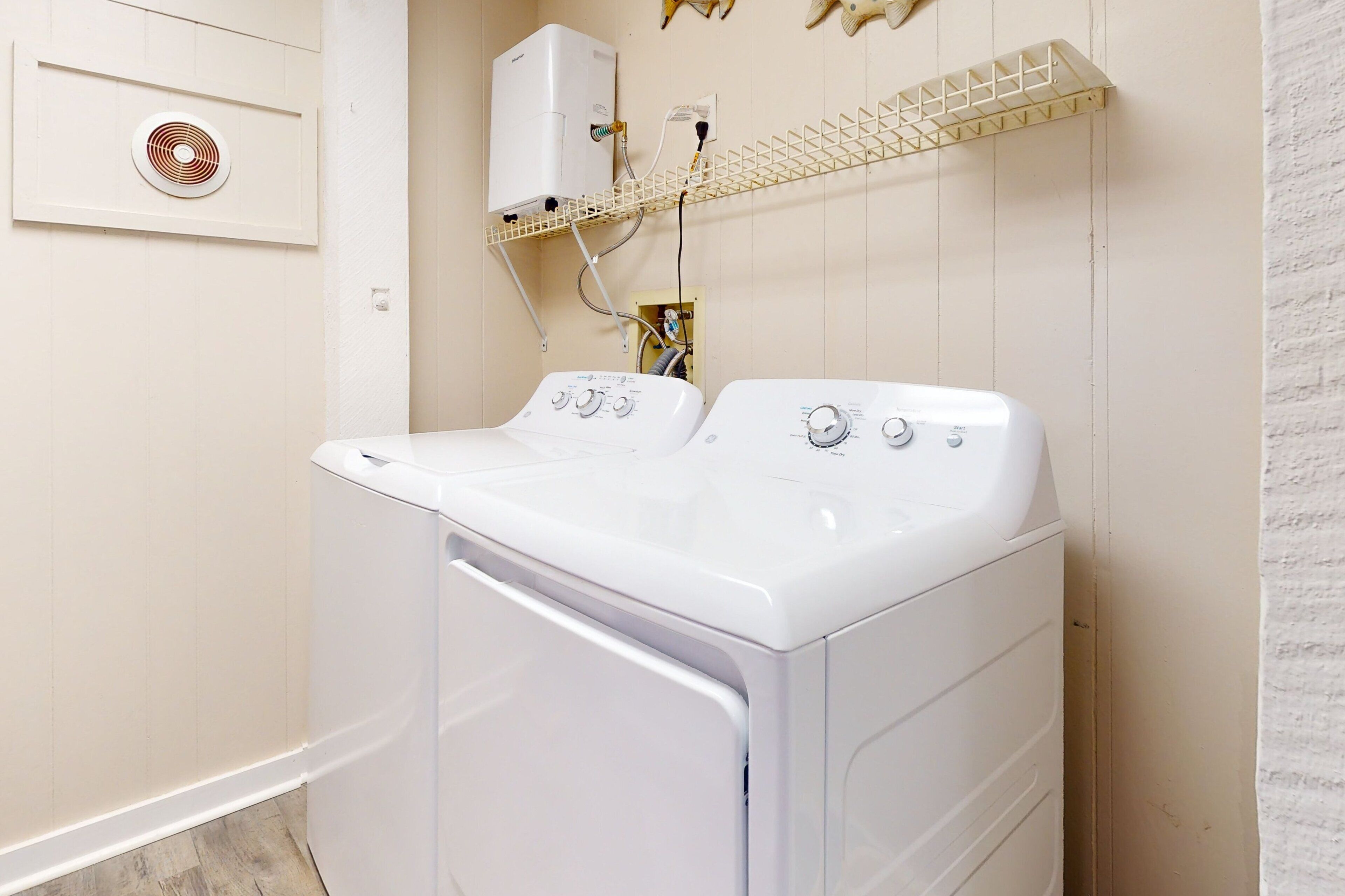 A laundry room with two white front-loading washing machines, a wall-mounted heater, and a shelf with hooks.