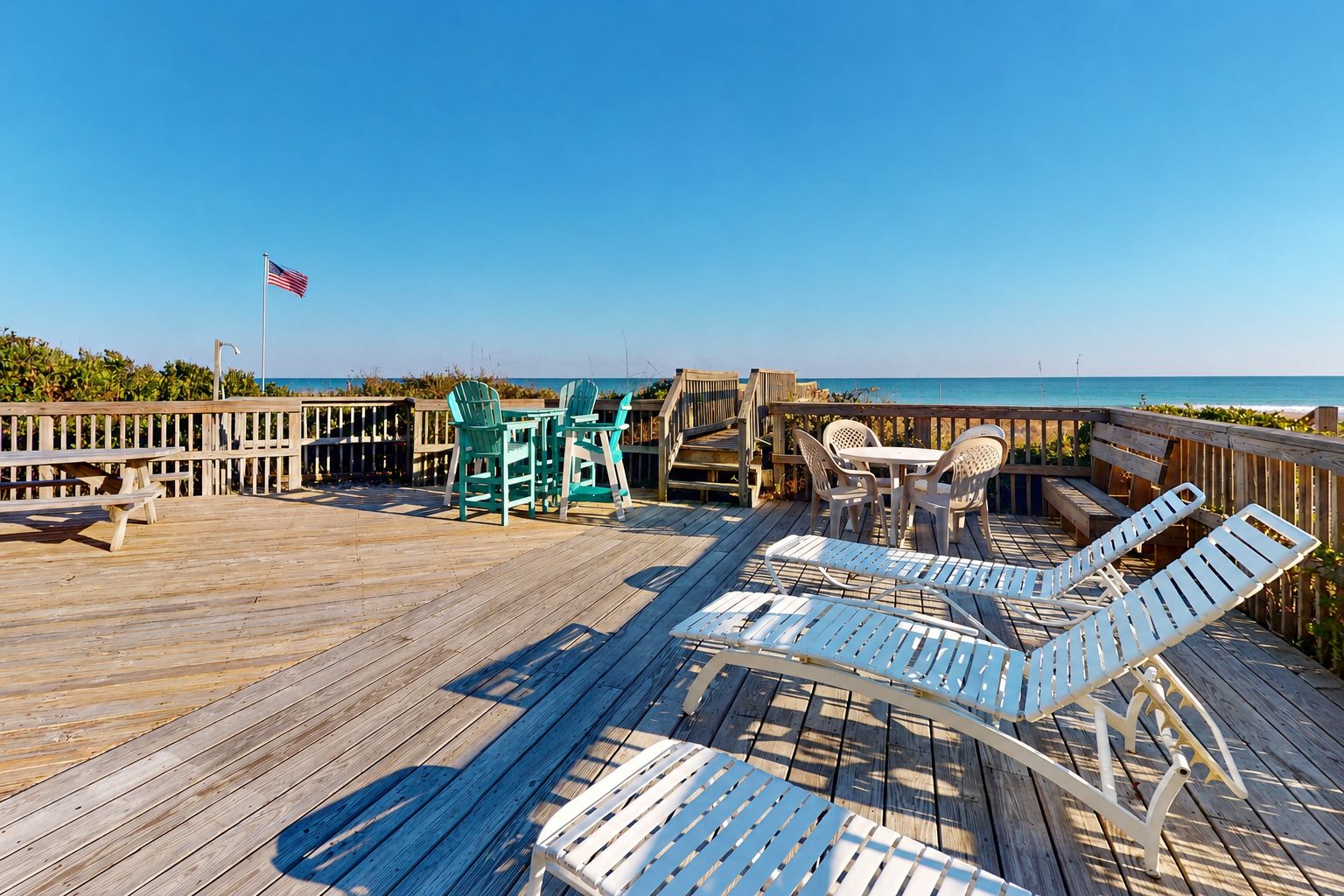 Eine Holzterrasse mit Sitzgelegenheiten im Freien, darunter Stühle und ein Tisch, mit Blick auf einen Strand.