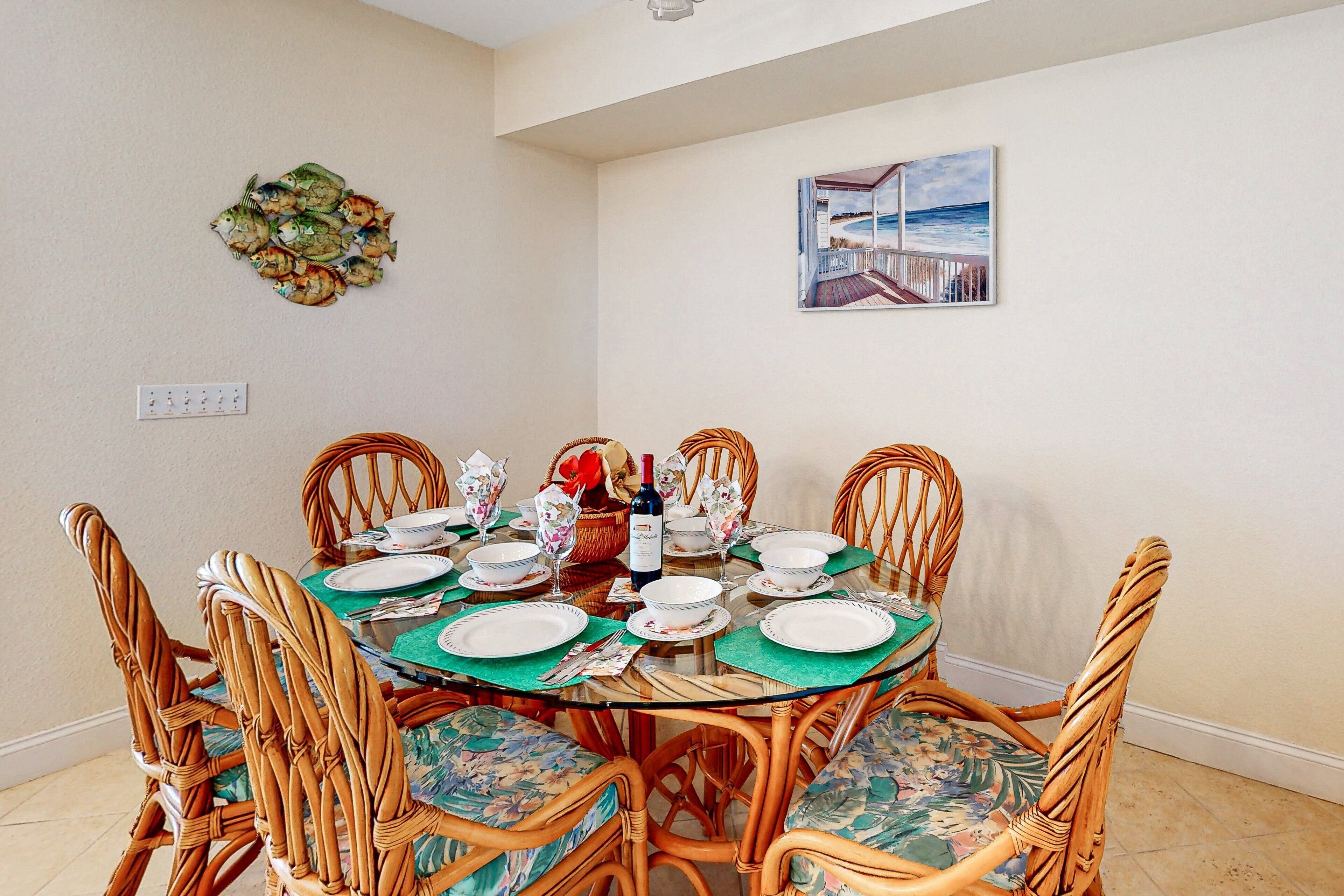 A dining area with a round glass table set for six, surrounded by wicker chairs with floral upholstery.