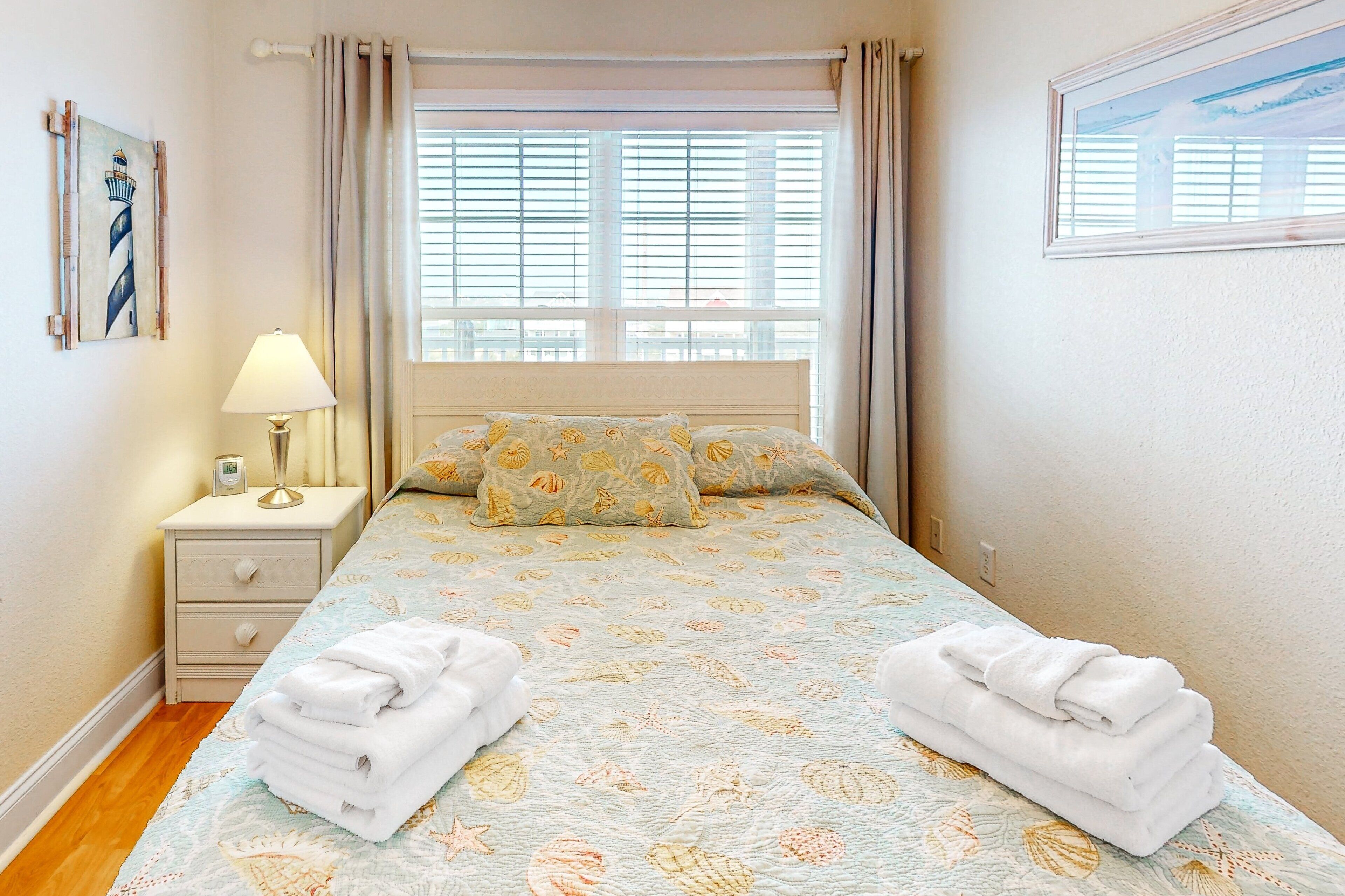 A neatly made bed with a light blue bedspread featuring a seashell pattern, two pillows with a similar design, a bedside table with a lamp, and a stack of folded towels.