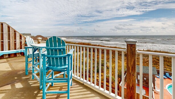 Un terrazzo in legno con poltrone Adirondack blu e un parapetto bianco con vista sulla spiaggia.