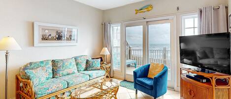 A living room with a sofa, armchair, TV, and a balcony with a view of the sea.