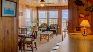 A wooden interior with a dining area, two sofas, a table, and a view through large windows. - 3BR Oceanfront Topsail Beach Cottage | Dog Friendly! (Topsail Beach)