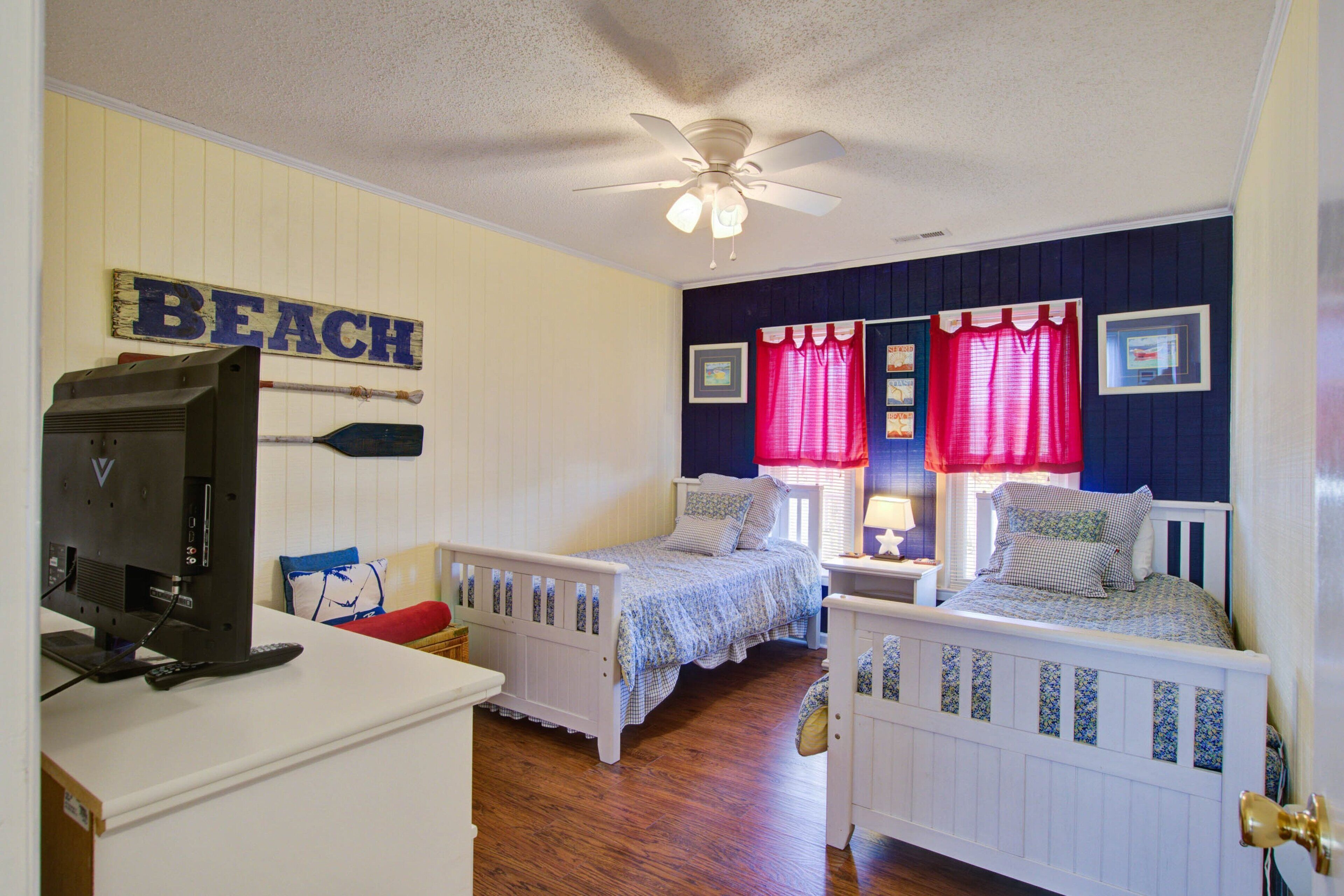 A bedroom with two beds, a ceiling fan, a TV, and a sign that reads "BEACH".