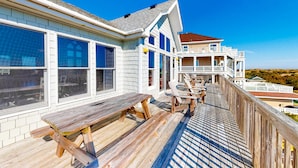 Eine Holzterrasse mit Picknicktischen und -stühlen, mit Blick auf einen Strand und ein großes Haus mit mehreren Balkonen.