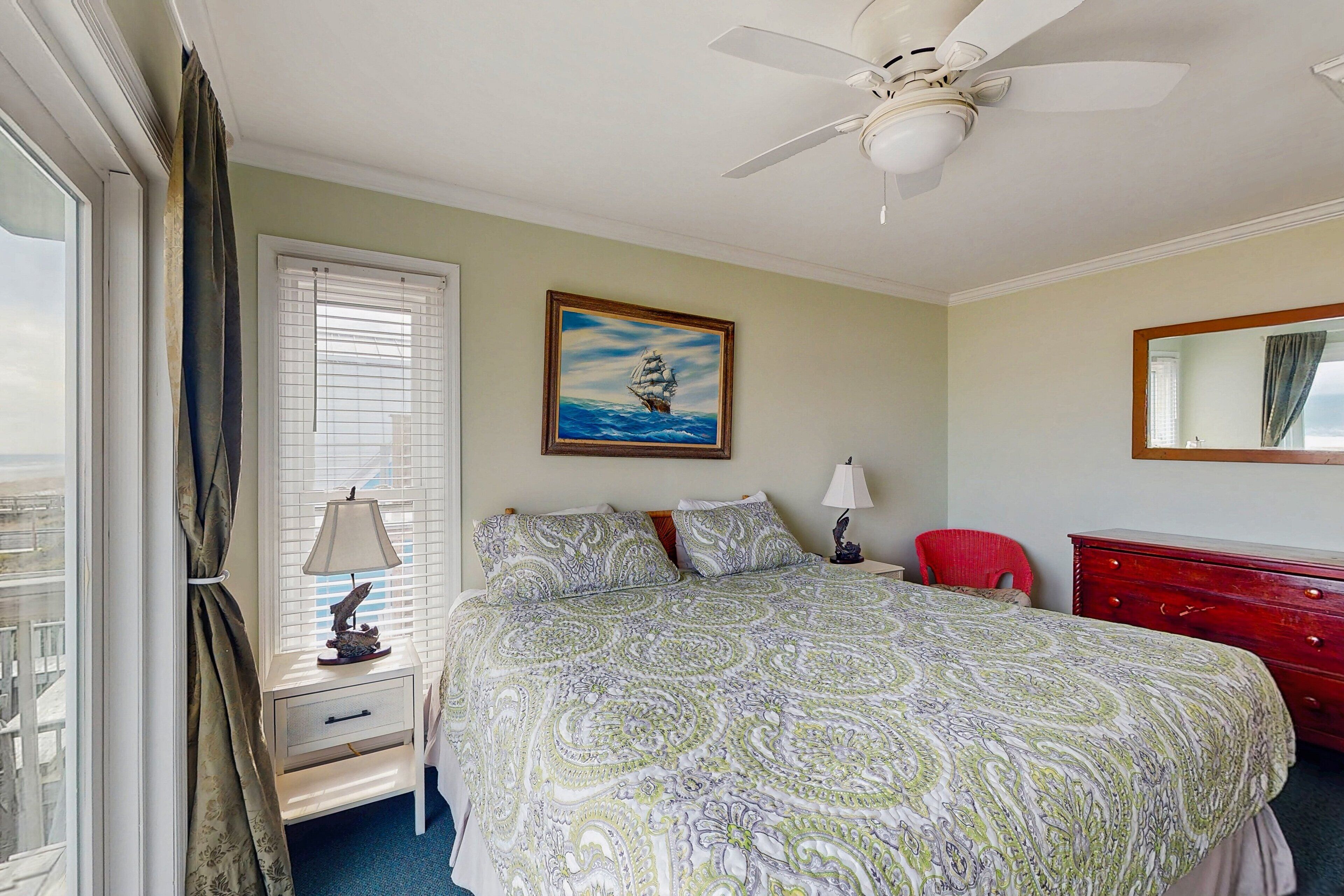A bedroom with a bed, a nightstand, a red chair, a dresser, a ceiling fan, and a painting of a ship on the wall.