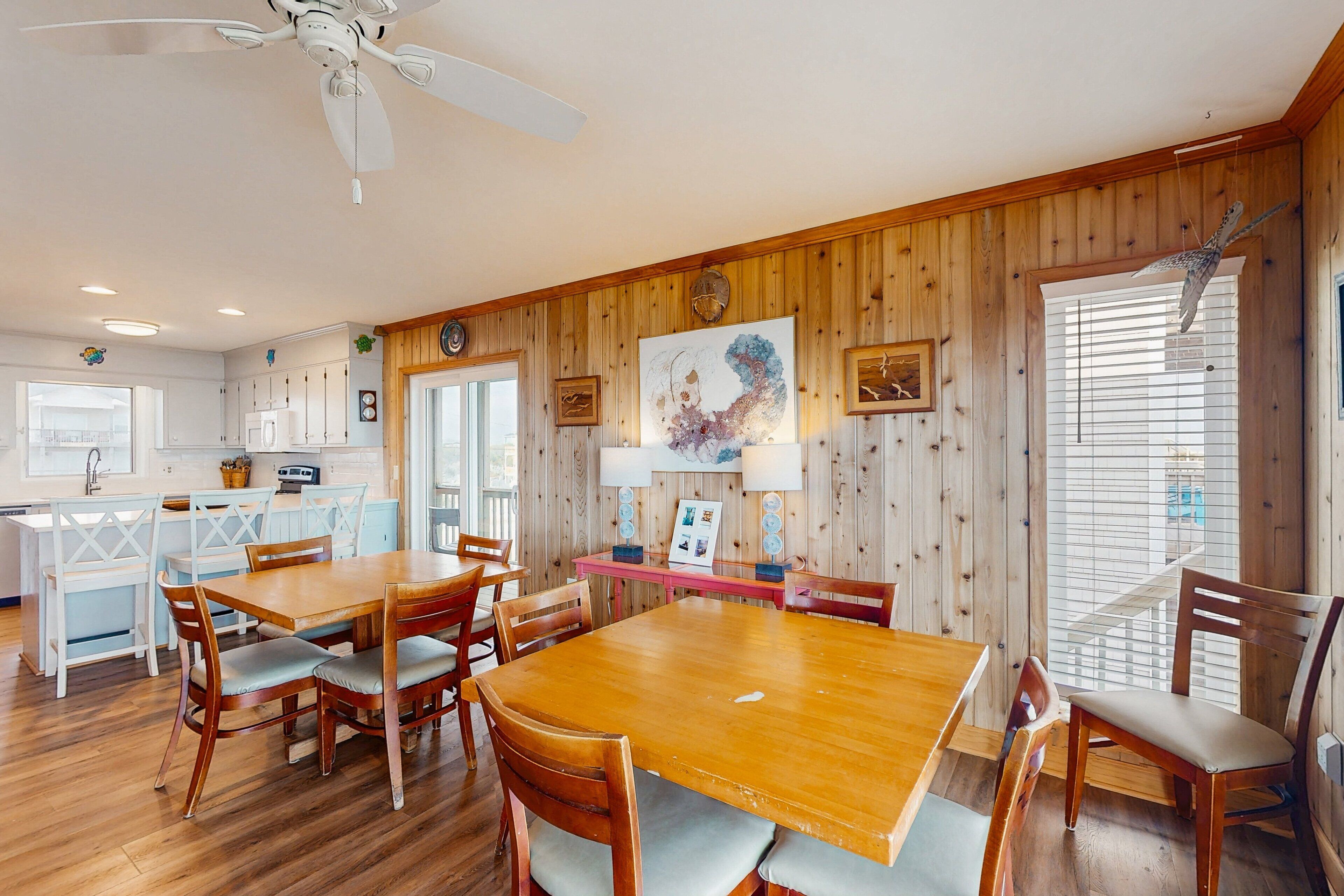 Un coin repas avec une table et des chaises en bois, un ventilateur de plafond et des murs en bois ornés d’œuvres d’art encadrées.