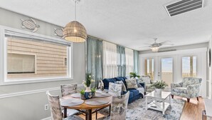 A spacious living room with a dining area, large windows, and a ceiling fan. - Lower Waterfront Condo with Oceanfront Balcony, Shared Outdoor Pool, WiFi & AC (Holden Beach)
