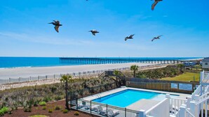 A pool area with a view of the beach and pier, surrounded by a fence and palm trees. - Lower Waterfront Condo with Oceanfront Balcony, Shared Outdoor Pool, WiFi & AC (Holden Beach)