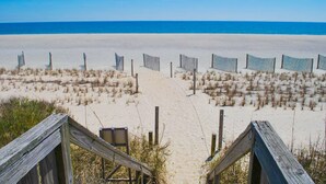 A beach with a wooden walkway leading to striped beach chairs and umbrellas. - Lower Waterfront Condo with Oceanfront Balcony, Shared Outdoor Pool, WiFi & AC (Holden Beach)