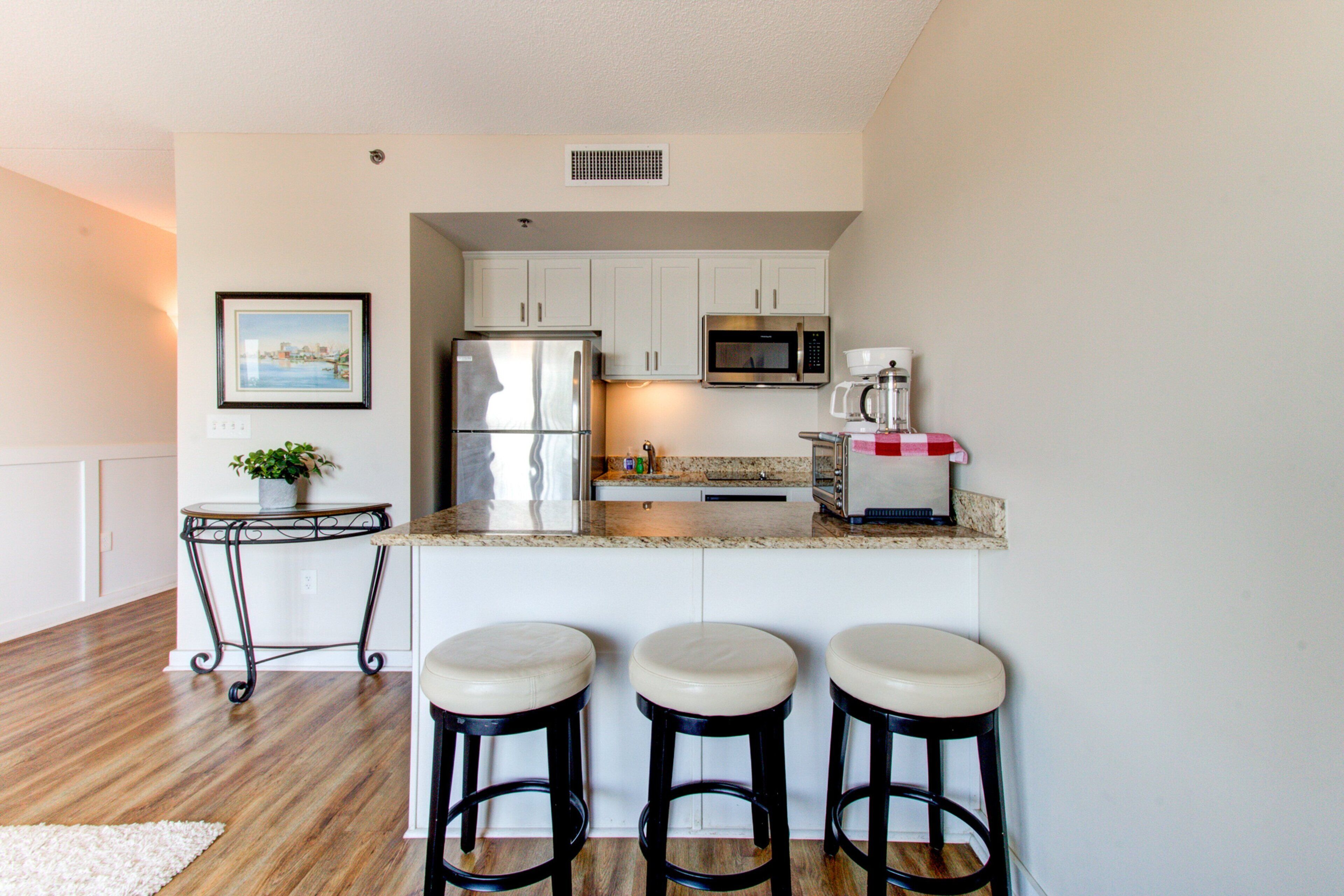 A modern kitchen with white cabinets, stainless steel appliances, and a granite countertop. There are three bar stools and a small table with a plant.