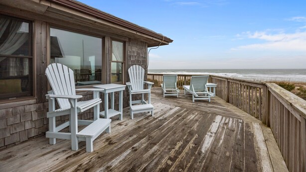 Eine Holzterrasse mit Adirondack-Stühlen und einem Tisch mit Blick auf den Ozean.