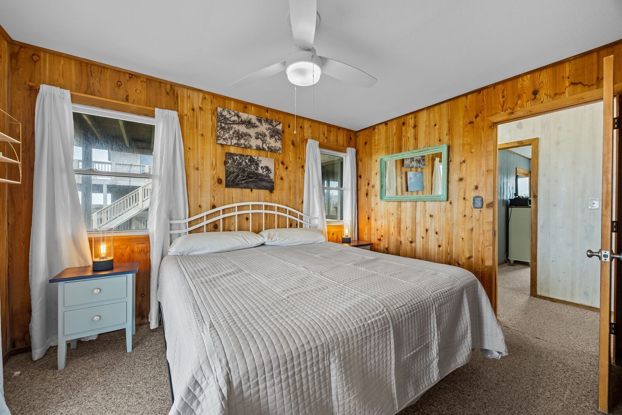 A bedroom with a wooden paneled wall, a white bed, a nightstand, a ceiling fan, and a window with curtains.