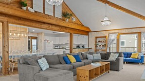 Ein großzügiges Wohnzimmer mit einem grauen Ecksofa, einem Holztisch und einem blauen Sofa. Im Hintergrund ist eine Küchenzeile zu sehen.