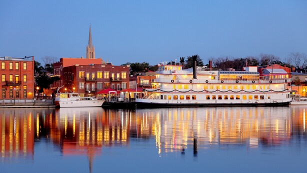 A multi-story riverboat with multiple decks, docked at a waterfront with buildings in the background.