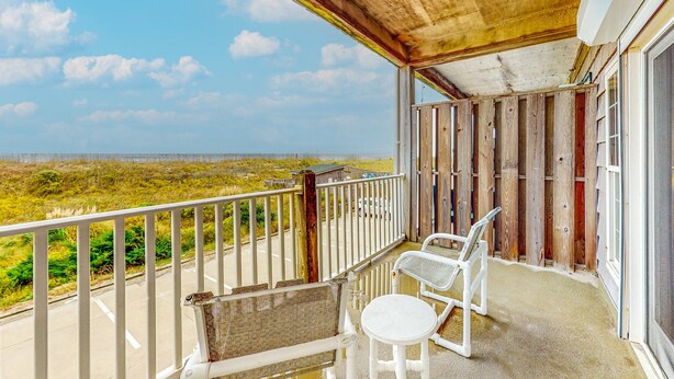 A balcony with a view of the beach, a white chair, a wicker chair, and a small table.