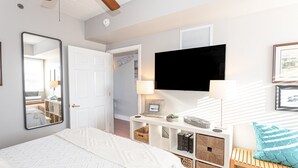 Une chambre avec un lit, un téléviseur fixé au mur, une table de chevet avec une lampe et un banc en bois avec un coussin.