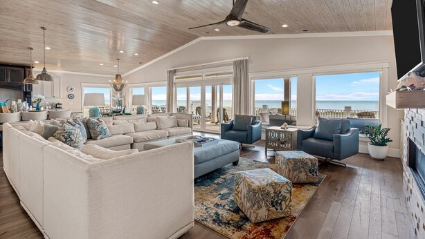 A spacious living room with a large sectional sofa, a blue armchair, and a coffee table. There is a ceiling fan, large windows with a view of the ocean, and a fireplace. - Modern Oceanfront Home with Zero-Entry Pool, Hot Tub, Outdoor Kitchen, WiFi & AC (Holden Beach)