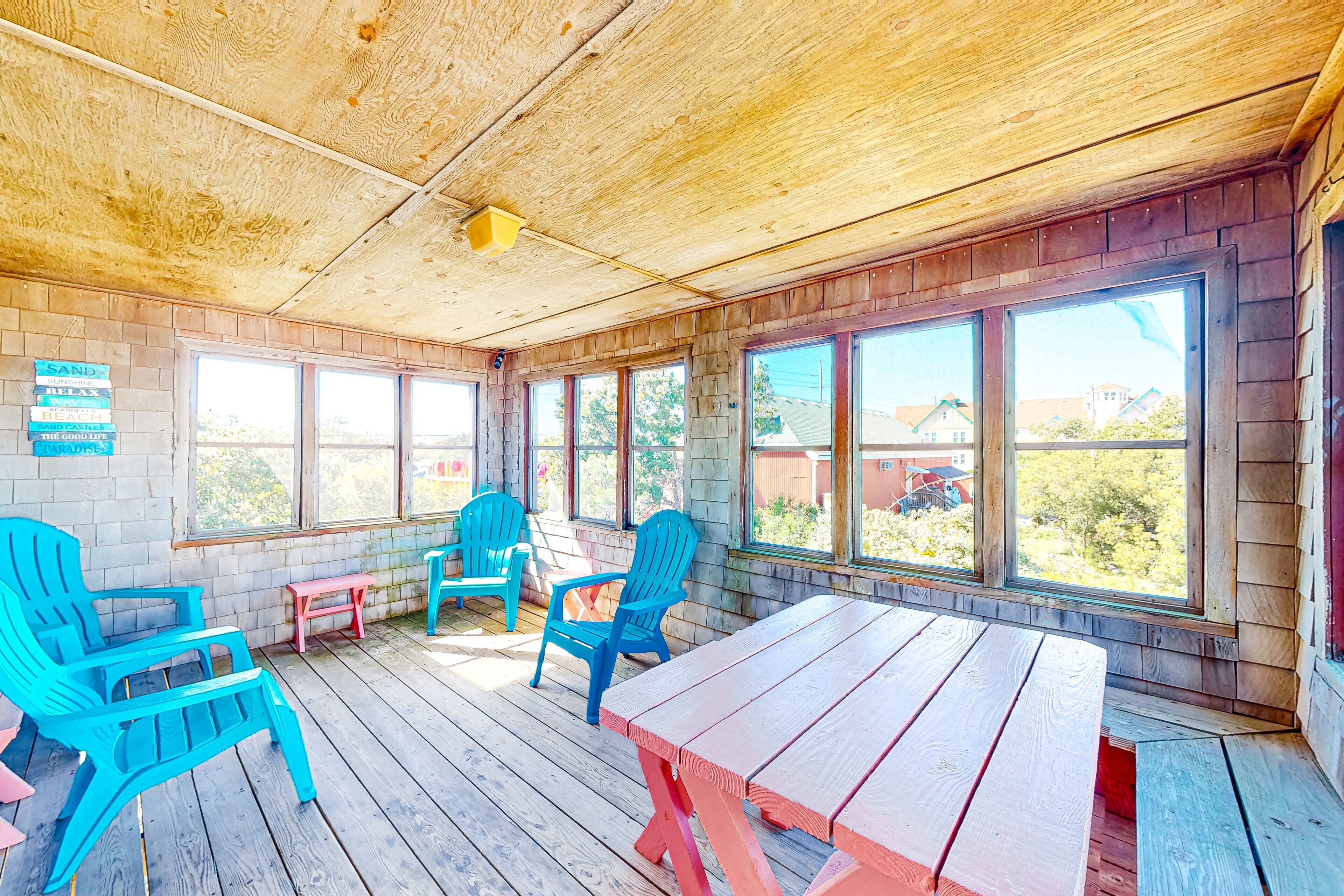 A covered patio with wooden flooring, a picnic table, and blue and pink chairs.