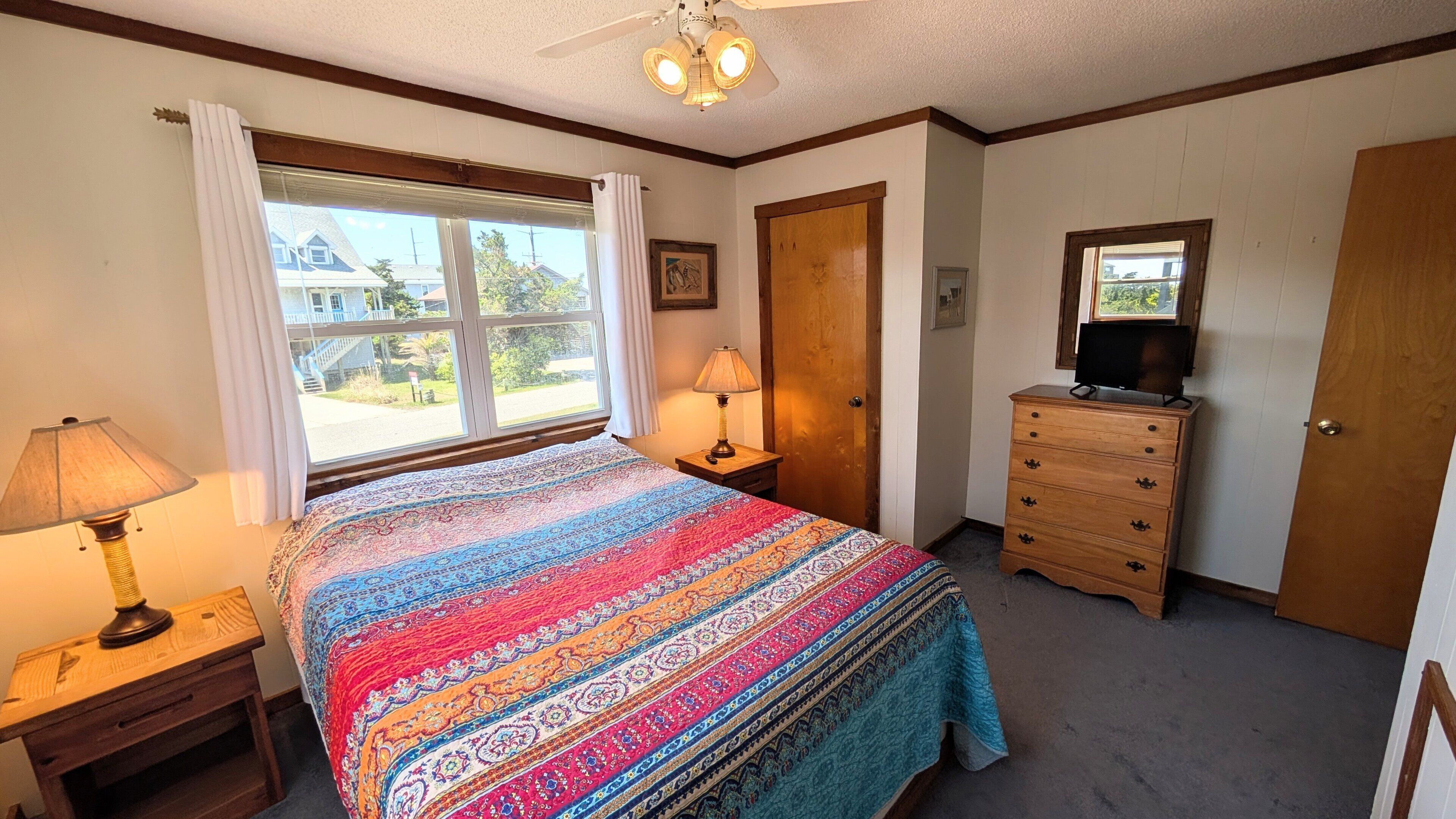 Une chambre à coucher avec un grand lit, deux tables de chevet avec des lampes, une commode, un ventilateur de plafond et une fenêtre avec des rideaux.