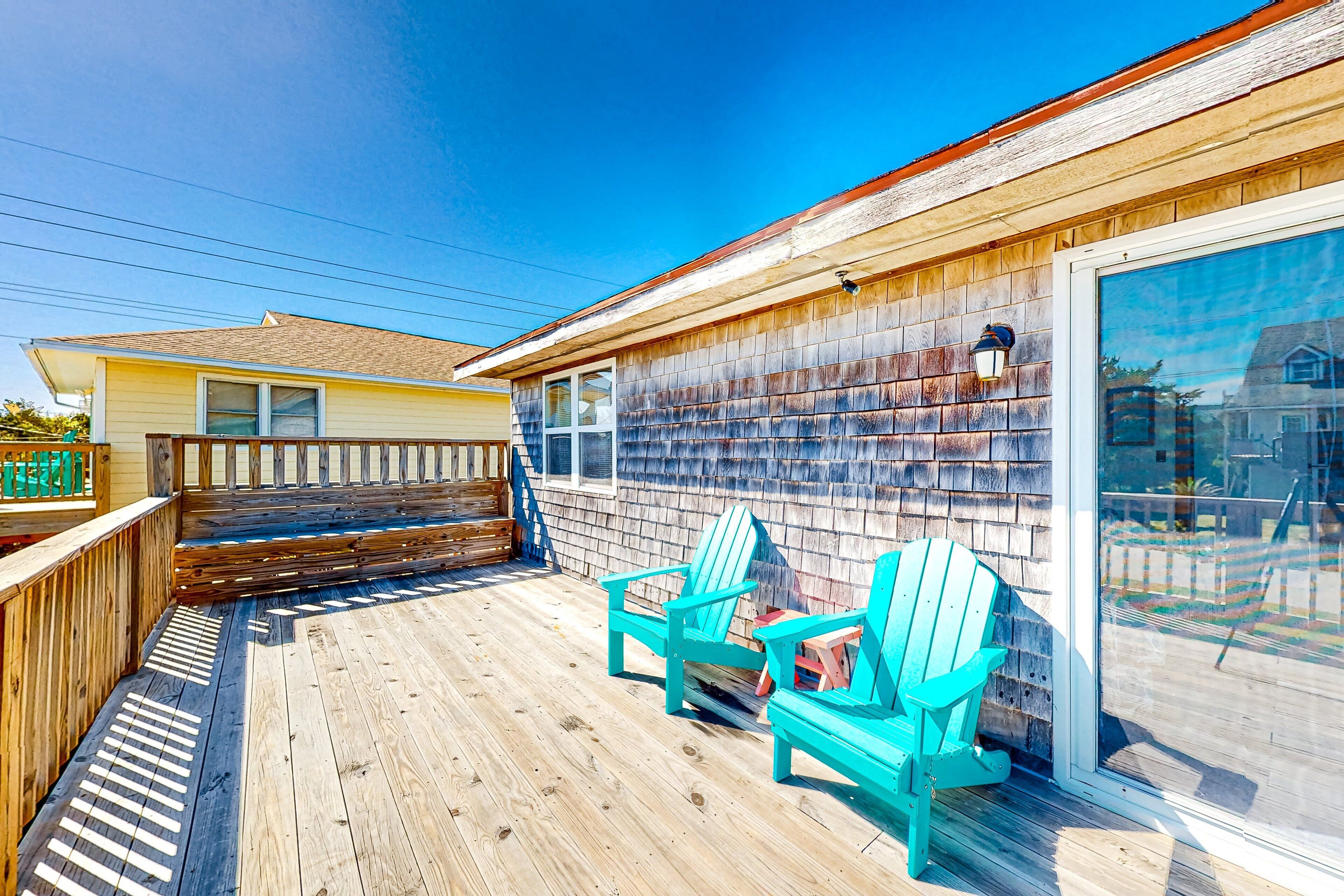 A wooden deck with two turquoise Adirondack chairs, a wooden bench, and a glass door leading to a covered area.