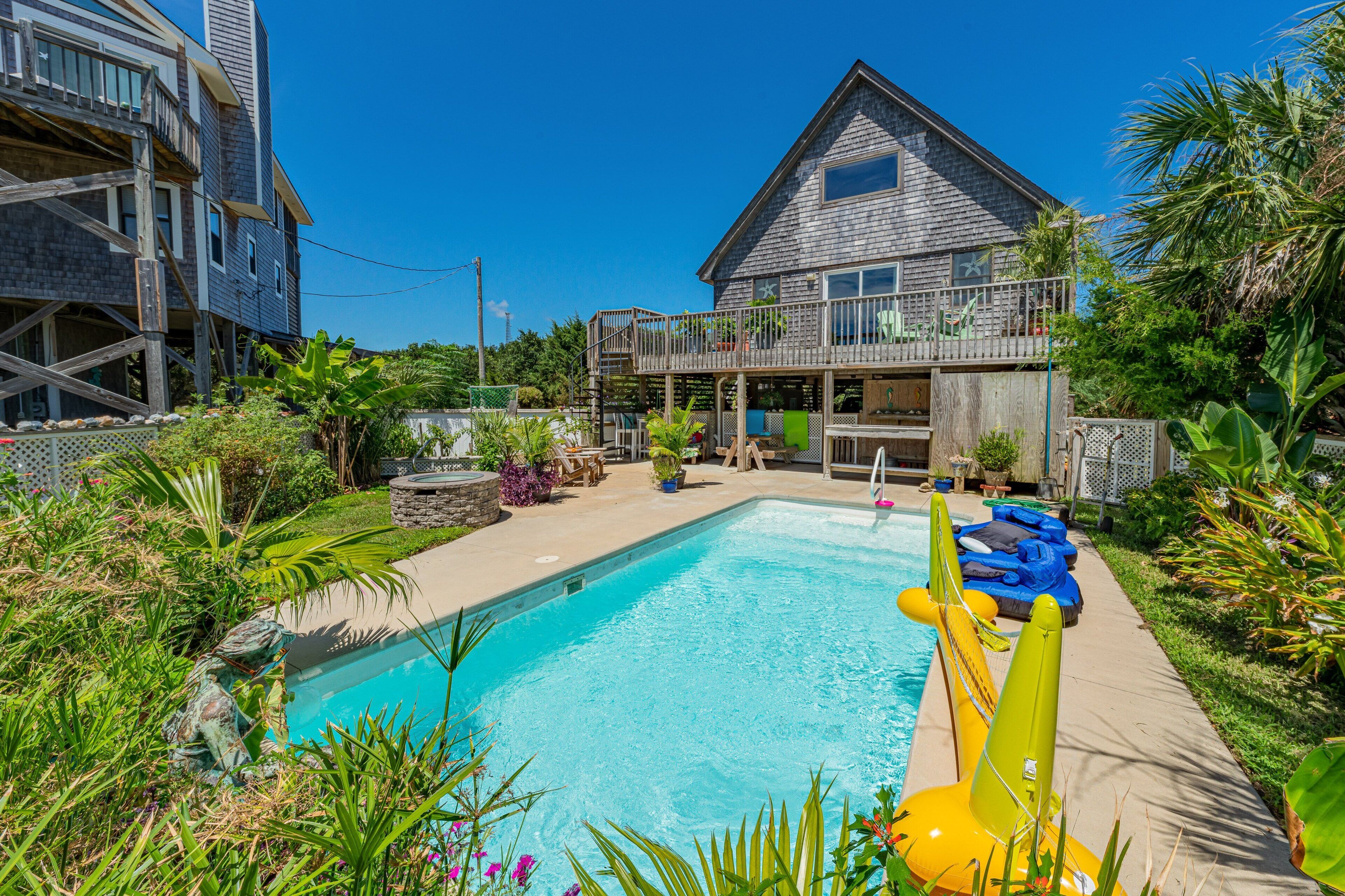 A swimming pool with a wooden deck, surrounded by lush greenery and a house with a balcony.