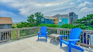 Une terrasse avec deux chaises Adirondack bleues et une petite table, d’où l’on a vue sur une plage et des maisons.