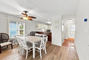 A dining area with a wooden table and white chairs, a ceiling fan, and a kitchen in the background. - Oceanfront home with WiFi, central AC, washer/dryer - dogs welcome (Holden Beach)