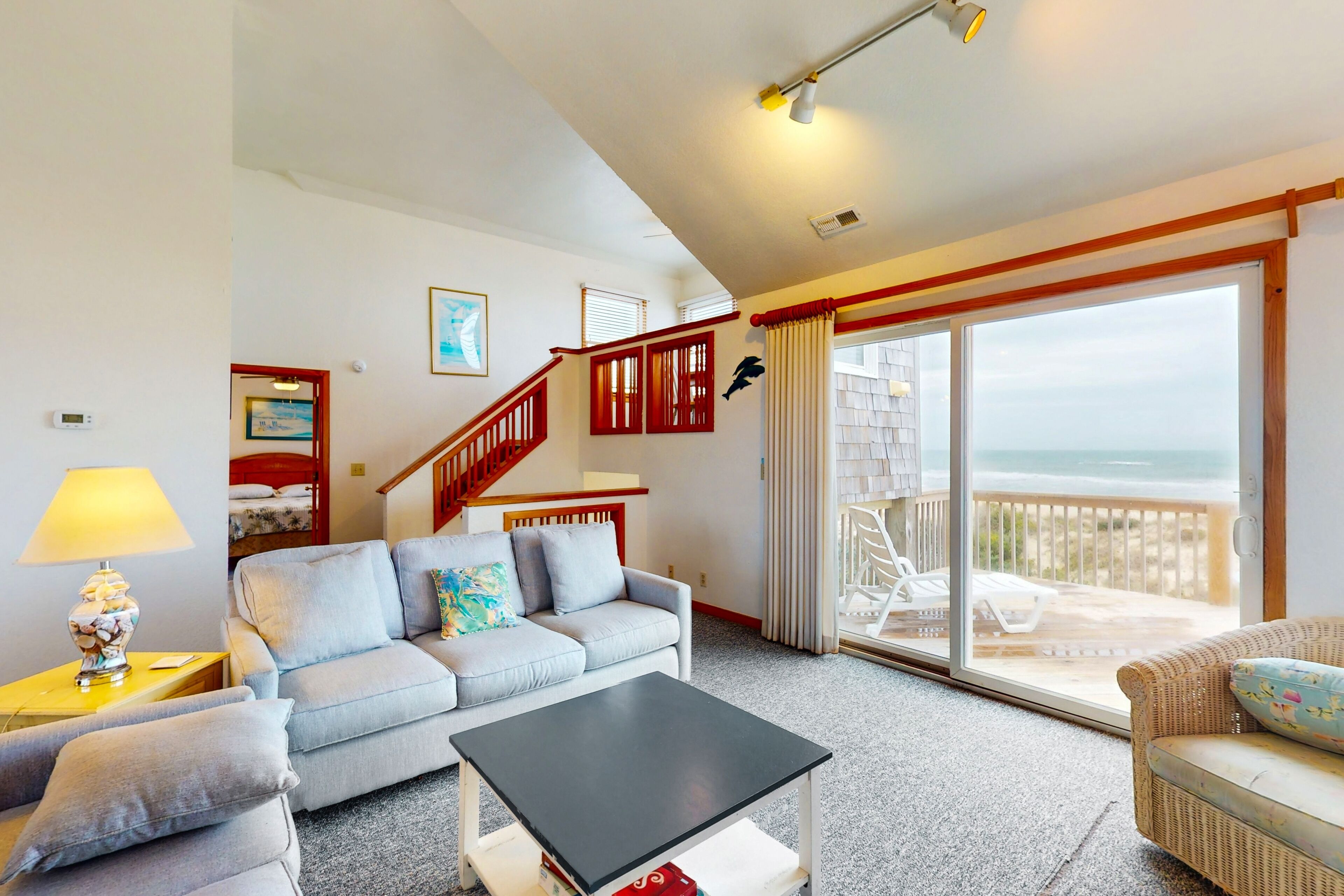 Uma sala de estar com sofá, mesa de centro, abajur, escada e varanda com vista para o mar.