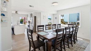 Cocina moderna con armarios blancos, una zona de comedor con mesa y sillas de madera, y un ventanal con vista a un edificio azul.