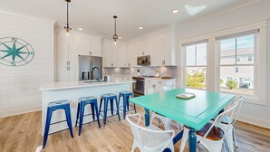 A modern kitchen with white cabinets, a central island with stools, and a teal dining table. - Of Course You Can: Private pool, patio, & balcony views (Holden Beach)