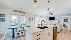 A modern kitchen with a central island, white cabinetry, and a dining area with chairs. - Of Course You Can: Private pool, patio, & balcony views (Holden Beach)