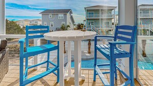 A covered outdoor seating area with blue chairs and a white table, overlooking a residential neighborhood with houses and a pool. - Of Course You Can: Private pool, patio, & balcony views (Holden Beach)