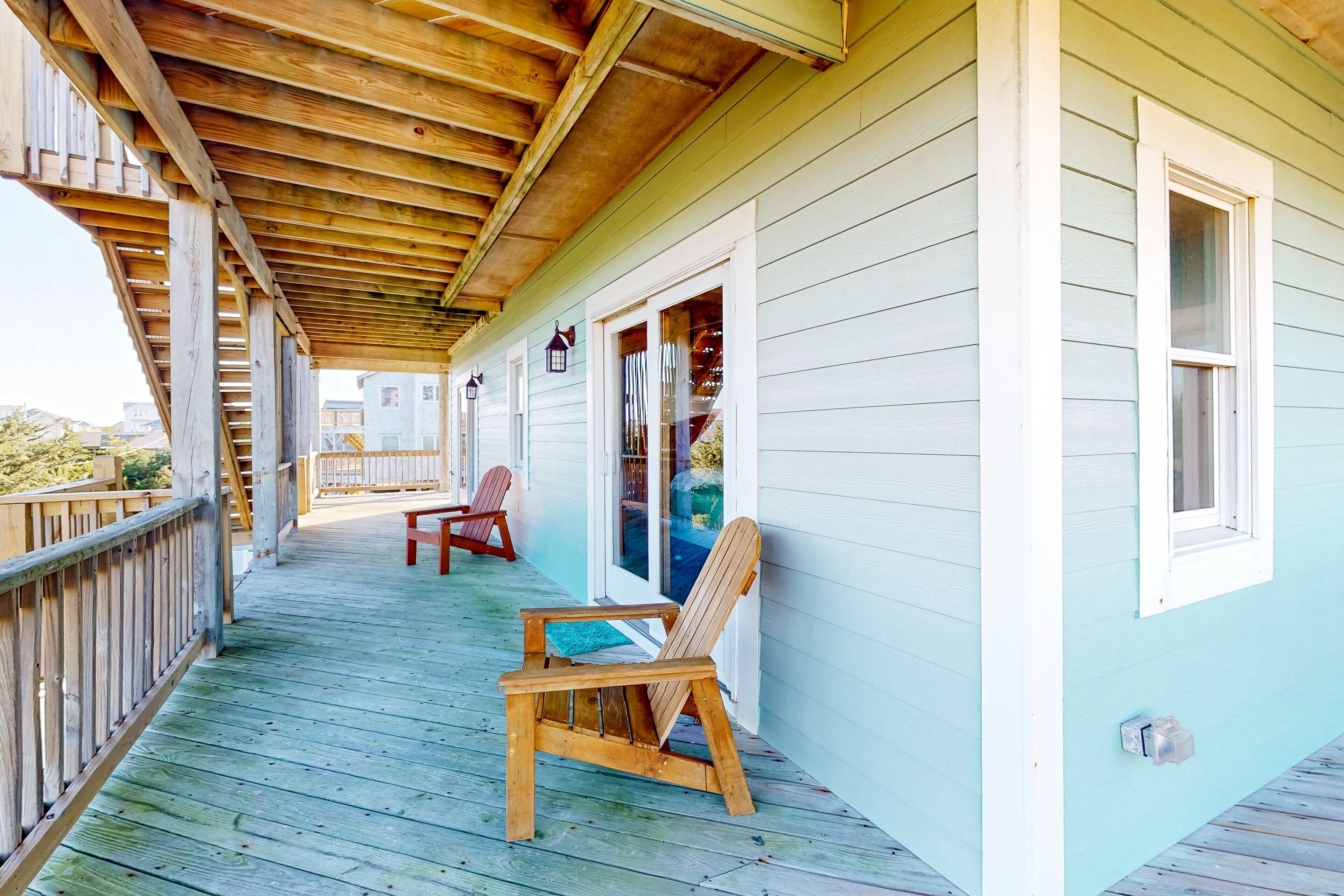 A wooden deck with two Adirondack chairs, a covered porch, and a light blue building with white trim.