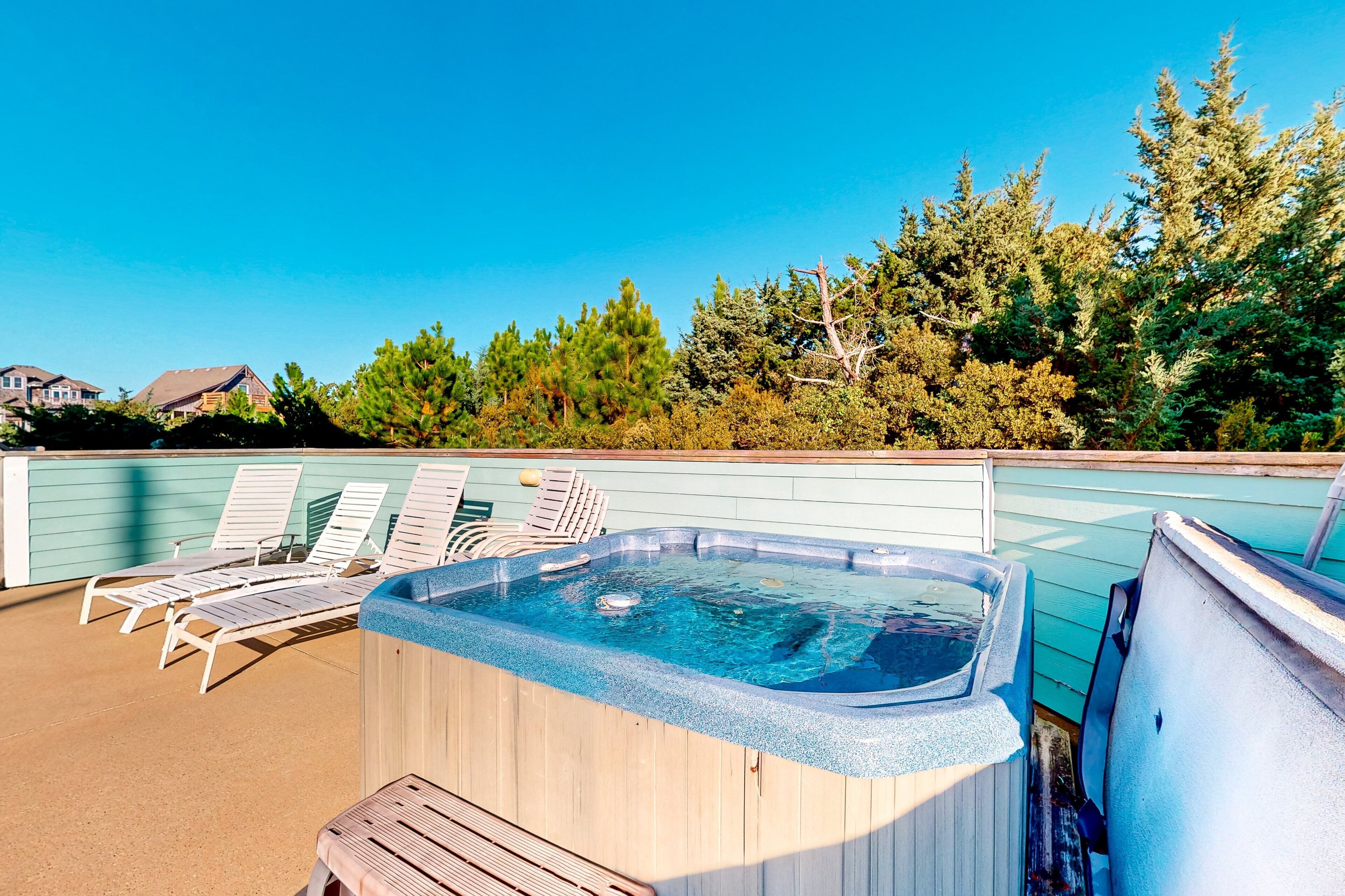 A hot tub on a patio with lounge chairs, surrounded by trees and a clear blue sky.