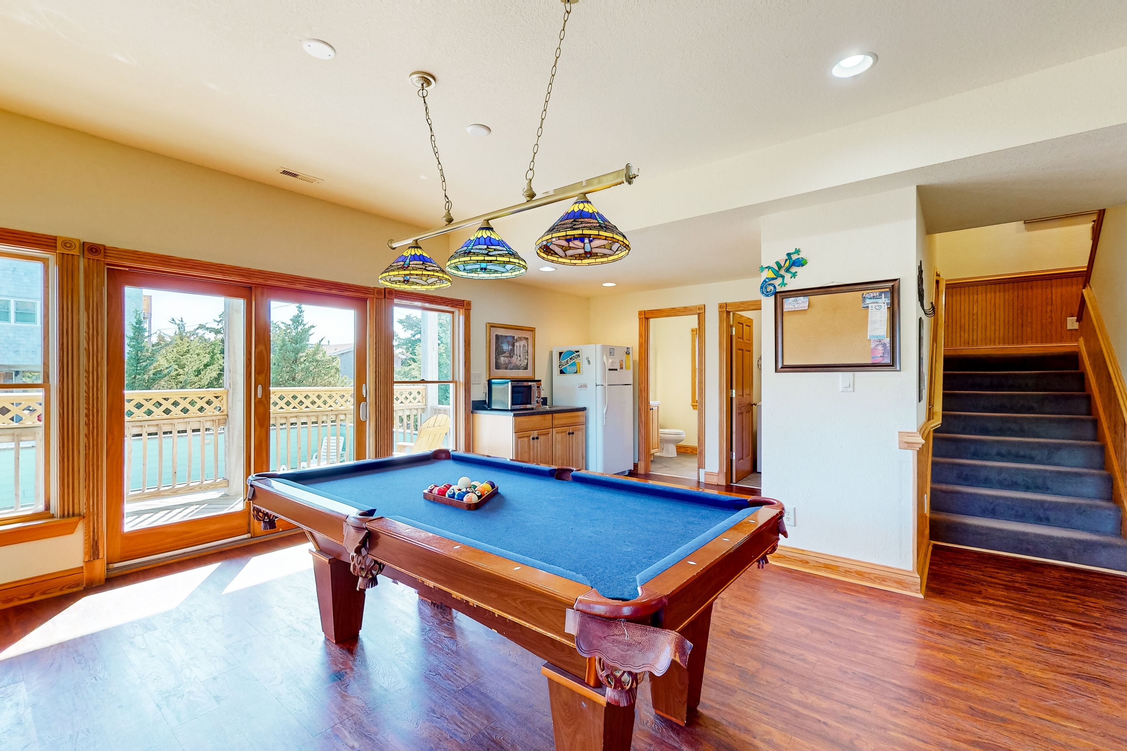 A pool table in a room with wooden flooring, a kitchen area, and a staircase.