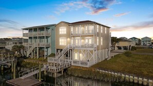A waterfront property with a two-story house, a dock, and a reflection in the water. - Turtle's Watch: Canal-front dog-friendly home near beach, canal view, & dock (Holden Beach)