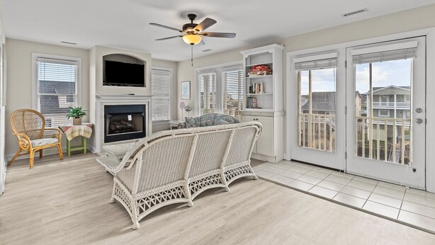 A spacious living room with a white wicker sofa, a fireplace, a ceiling fan, and a large window with blinds. - Turtle's Watch: Canal-front dog-friendly home near beach, canal view, & dock (Holden Beach)