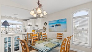 A dining area with a glass table surrounded by wooden chairs, white cabinets, and a painting of boats on the wall. - Turtle's Watch: Canal-front dog-friendly home near beach, canal view, & dock (Holden Beach)