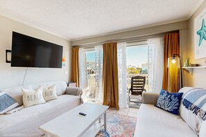 A hotel room with a flat-screen TV mounted on the wall, two sofas with decorative pillows, a small table, and a balcony with a view.