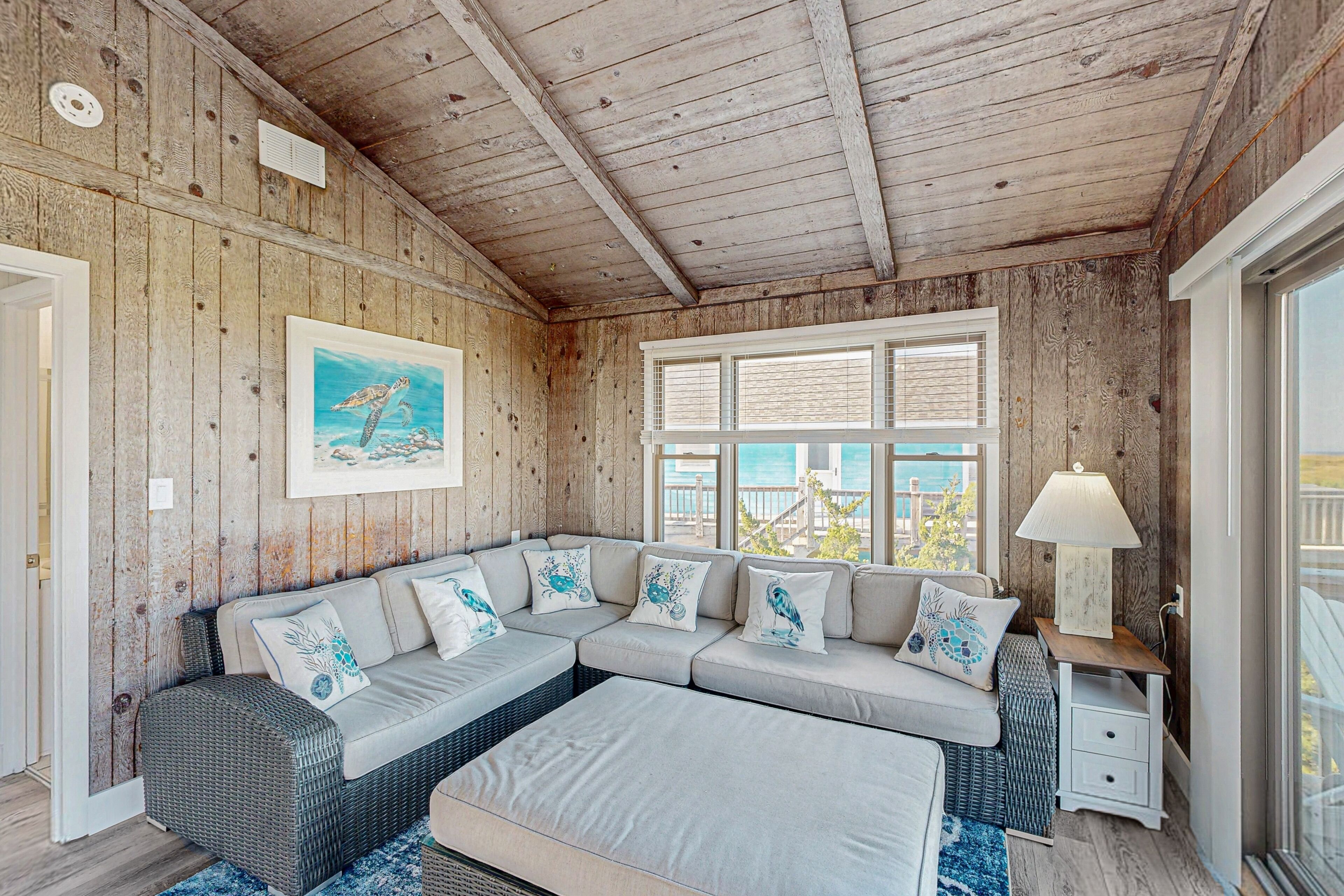 Una habitación acogedora con un sofá modular, techo de madera, un ventanal grande y una lámpara sobre una mesita.